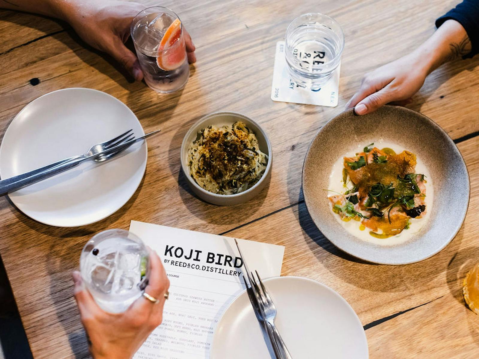 Food and drinks lay on a table in a restaurant, with hands holding some of them