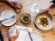 Food and drinks lay on a table in a restaurant, with hands holding some of them