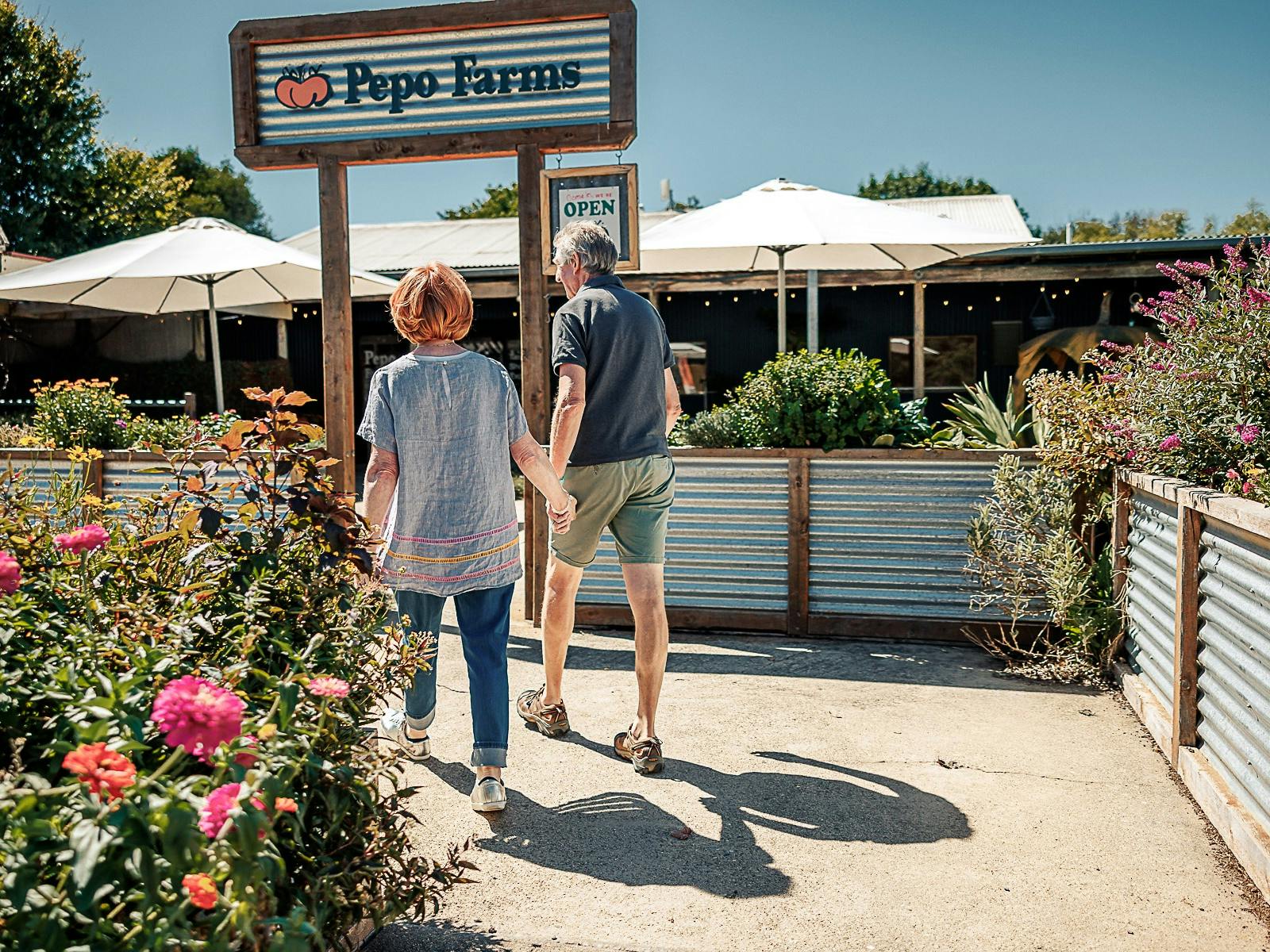 Couple walks hand-in-hand through garden toward Pepo Farms shopfront on a sunny day.
