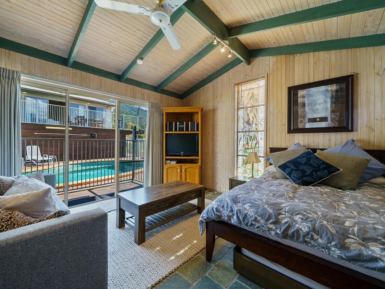 A pool house with a bed, couch and table. Swimming pool in the background outside.