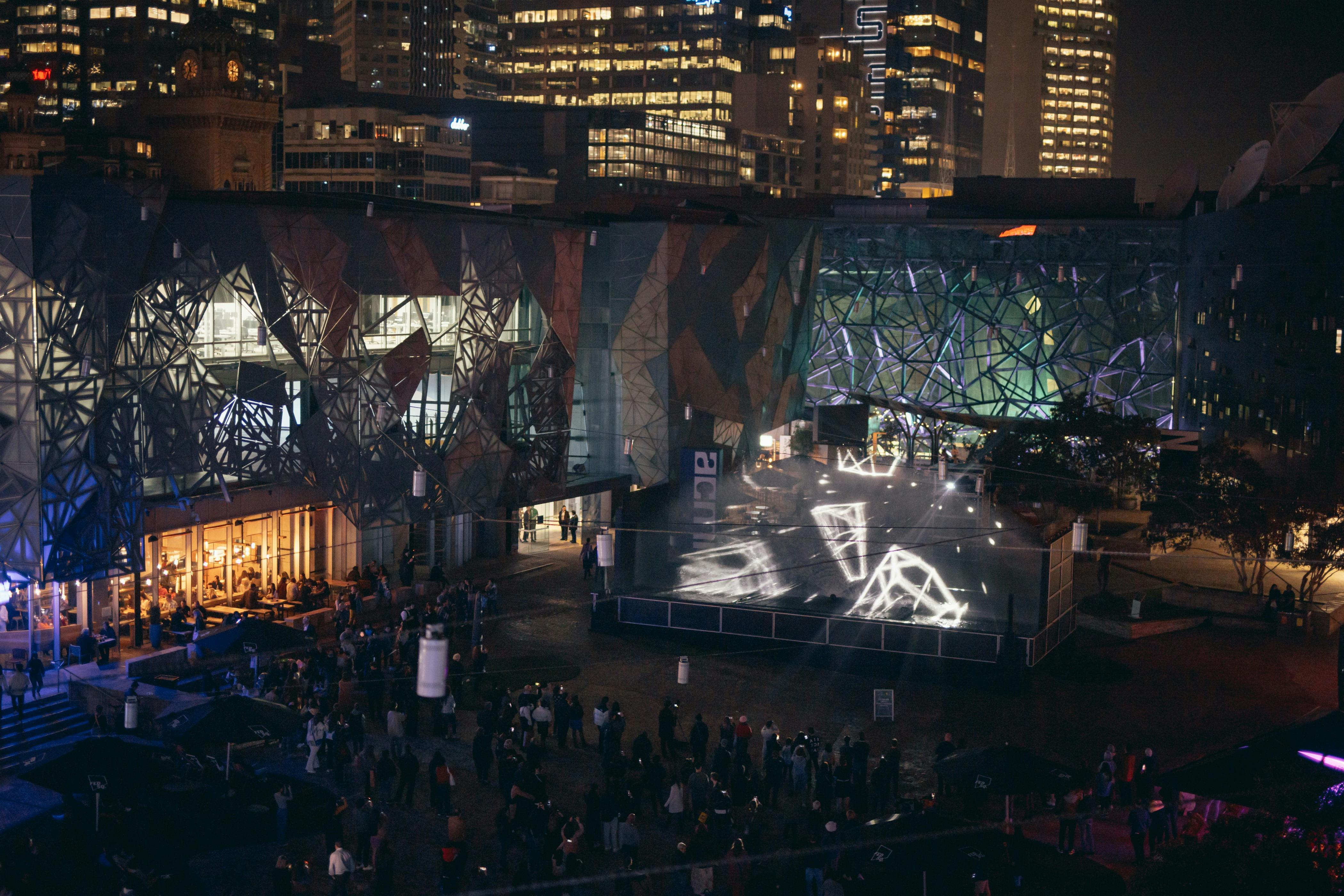 An overhead view of Fed Square at night, with people watching a laser light show.