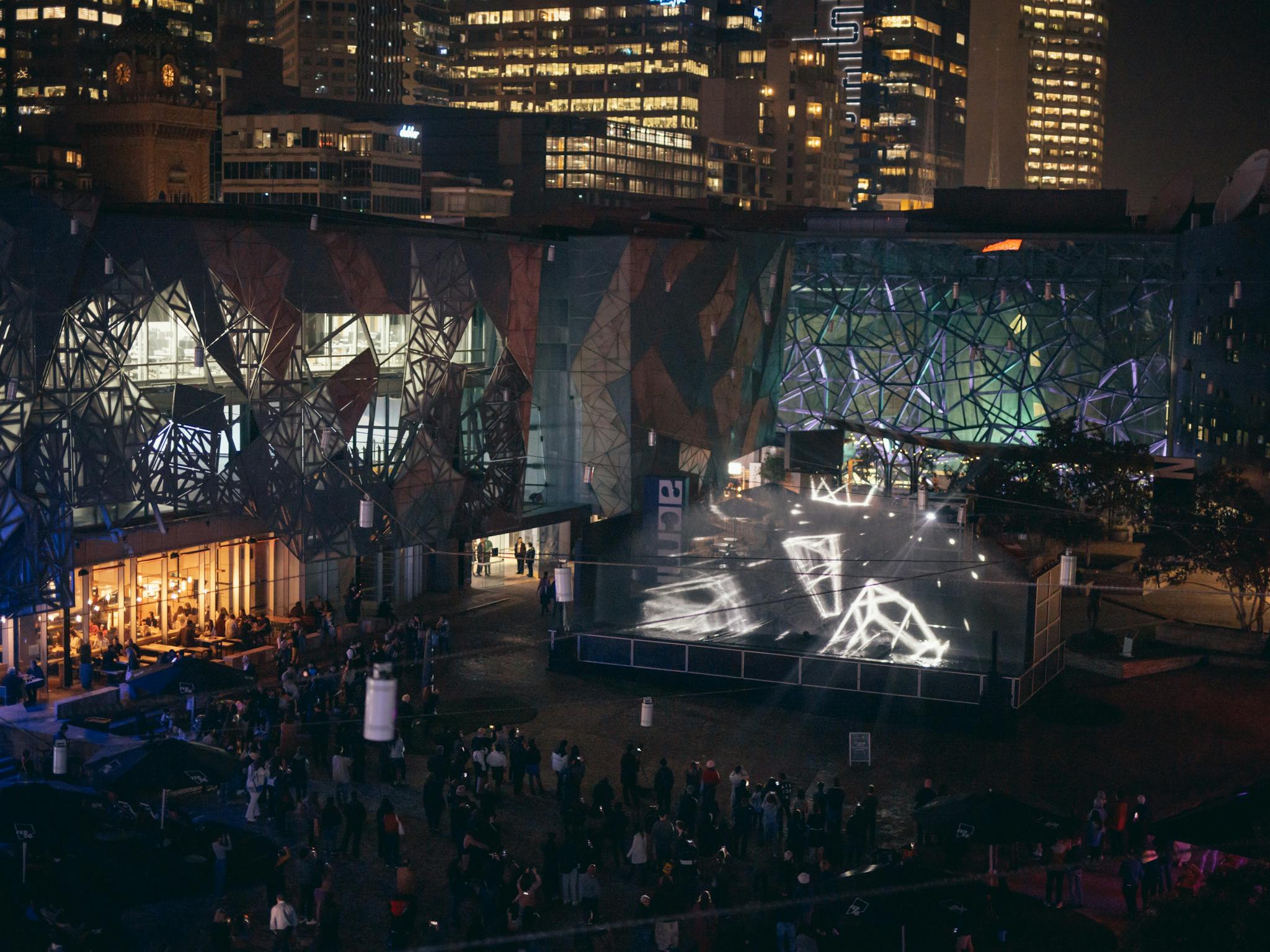 An overhead view of Fed Square at night, with people watching a laser light show.