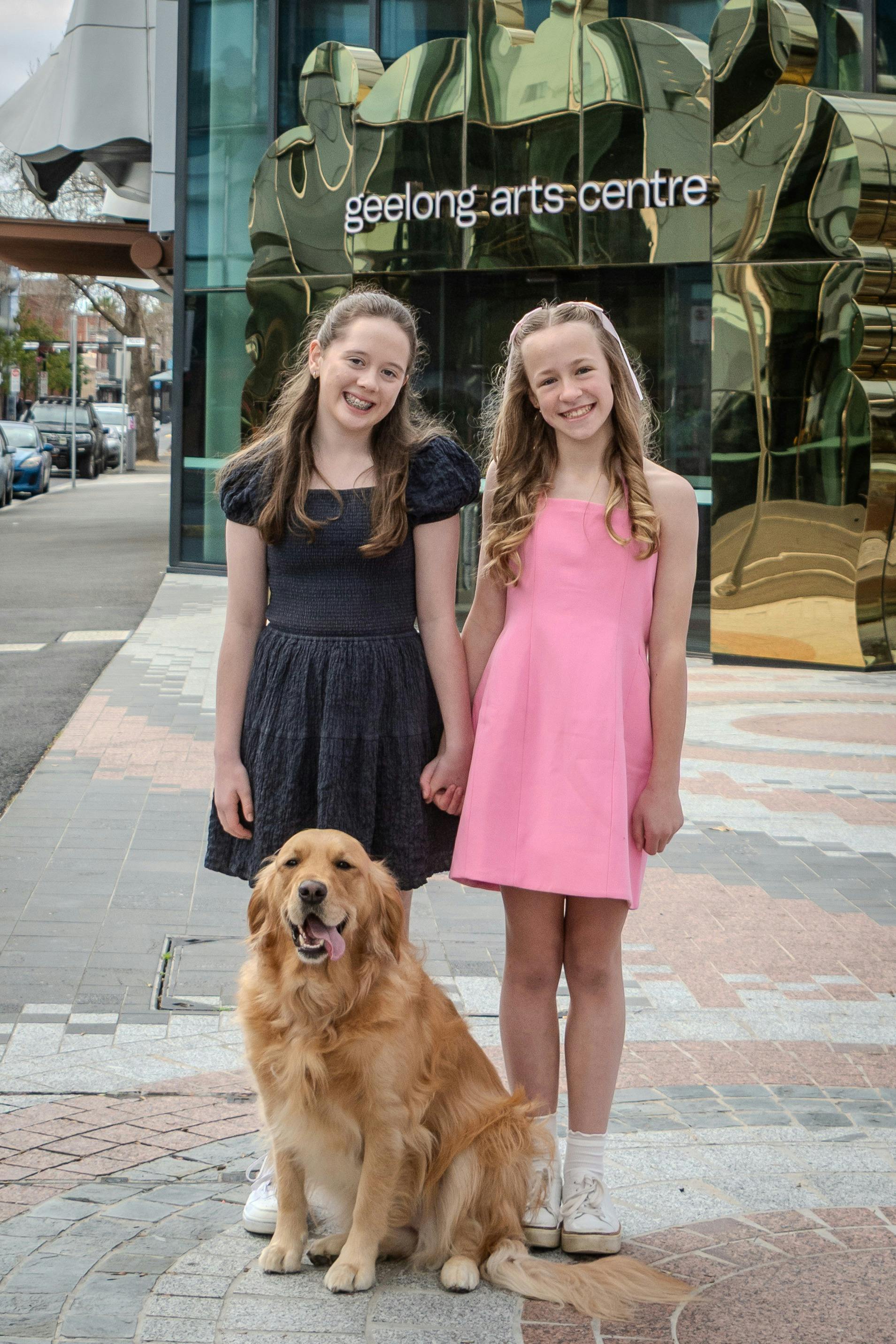 Bella McConachy and Clementine Oliver posing with a dog in front of Geelong Arts Centre's gold doors