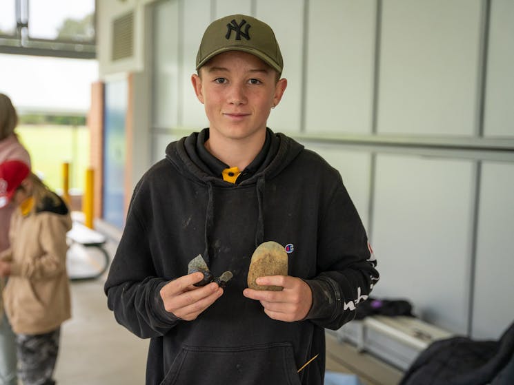 A young boy wearing a hat holding traditional Aboriginal stone tools.