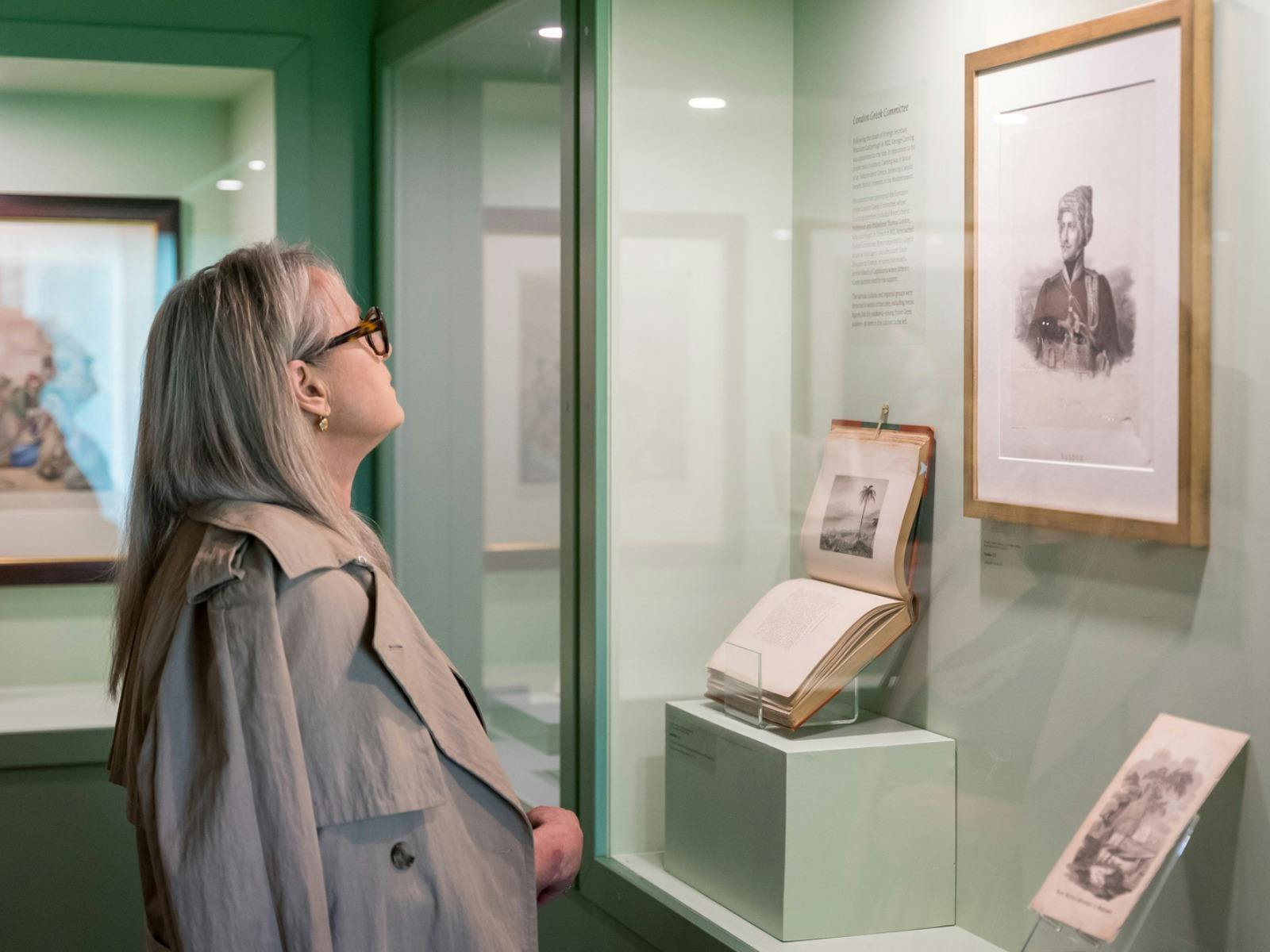 A blonde woman in a trench coat looks at a framed artwork and book display in a green gallery