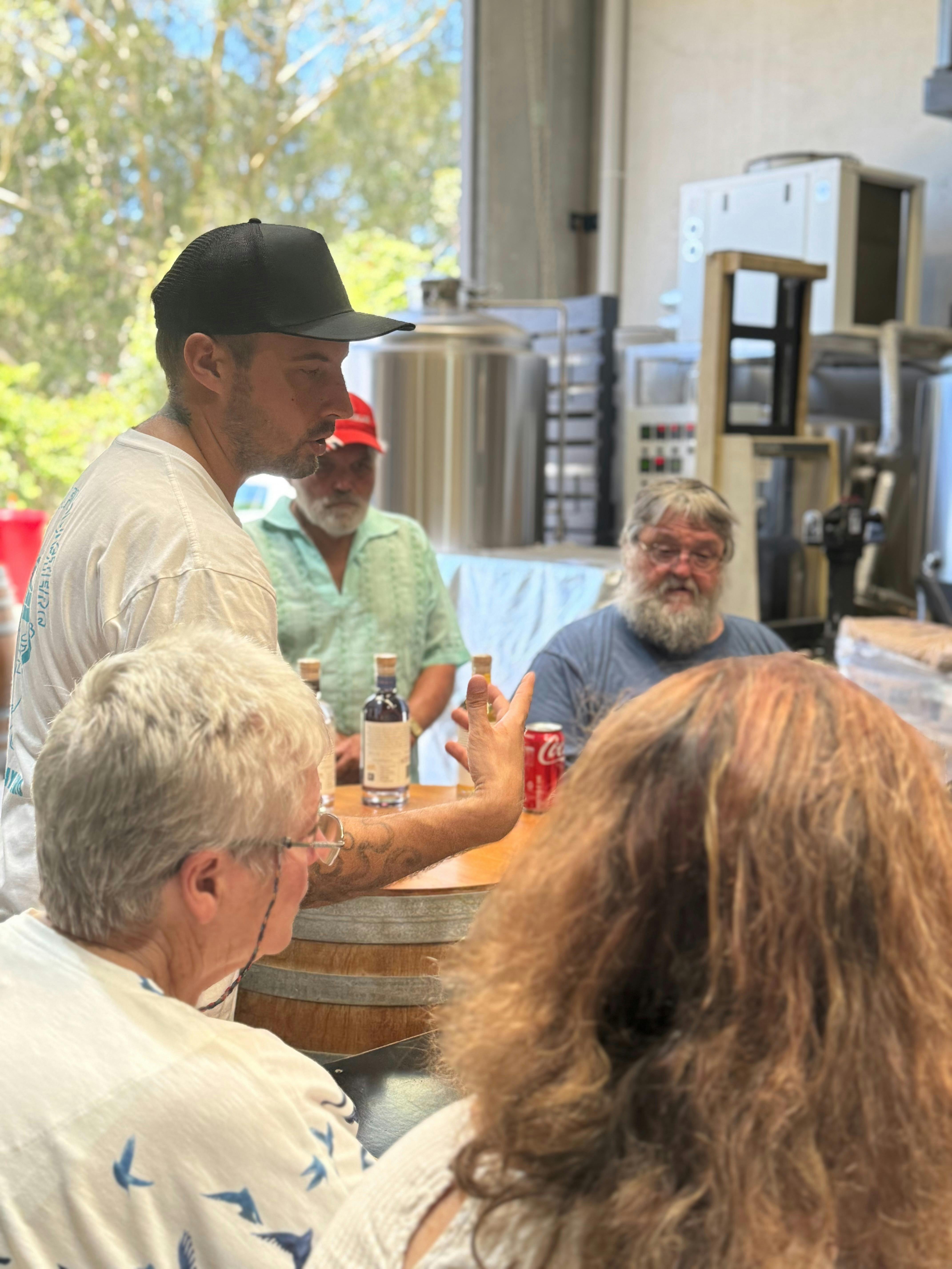 A brewery host speaks to a group gathered around a wooden table, with brewing equipment behind them.