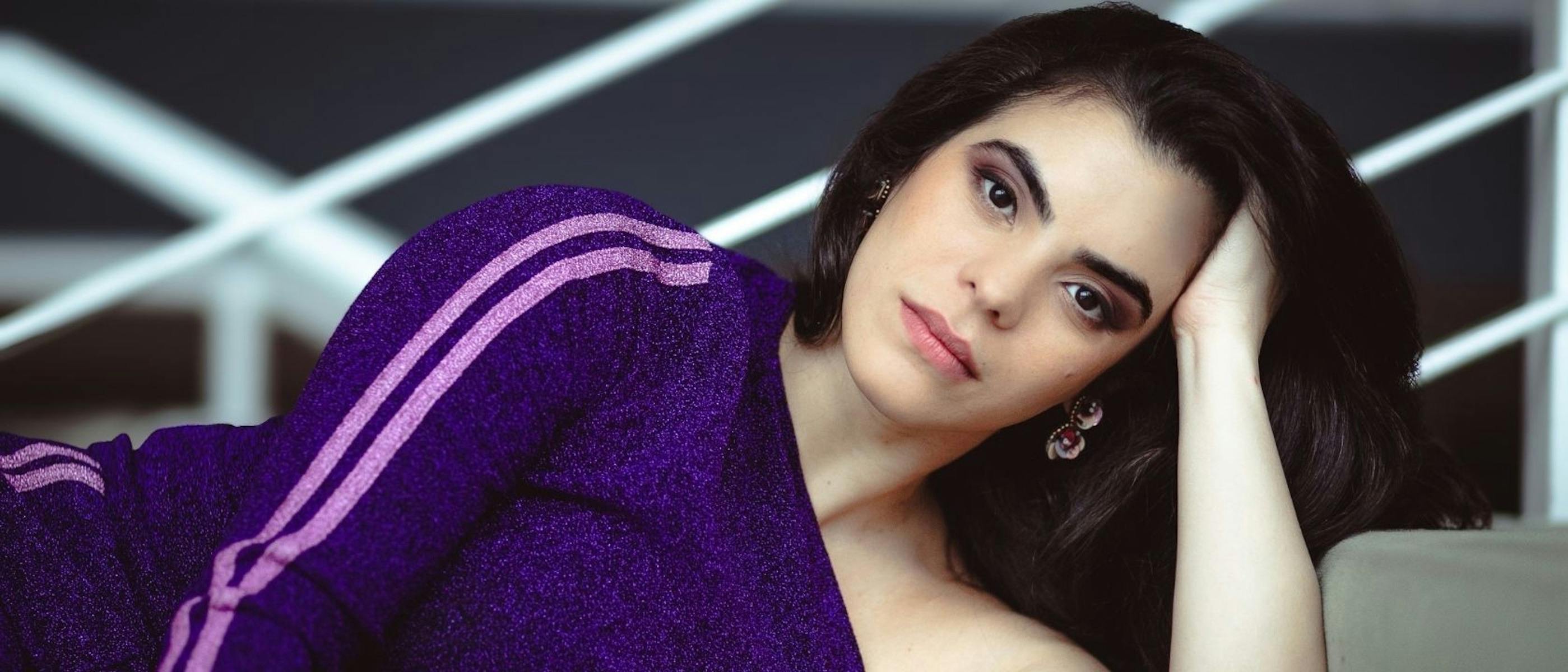 A close-up image of a woman with brown hair wearing a purple top, leaning on her hand.