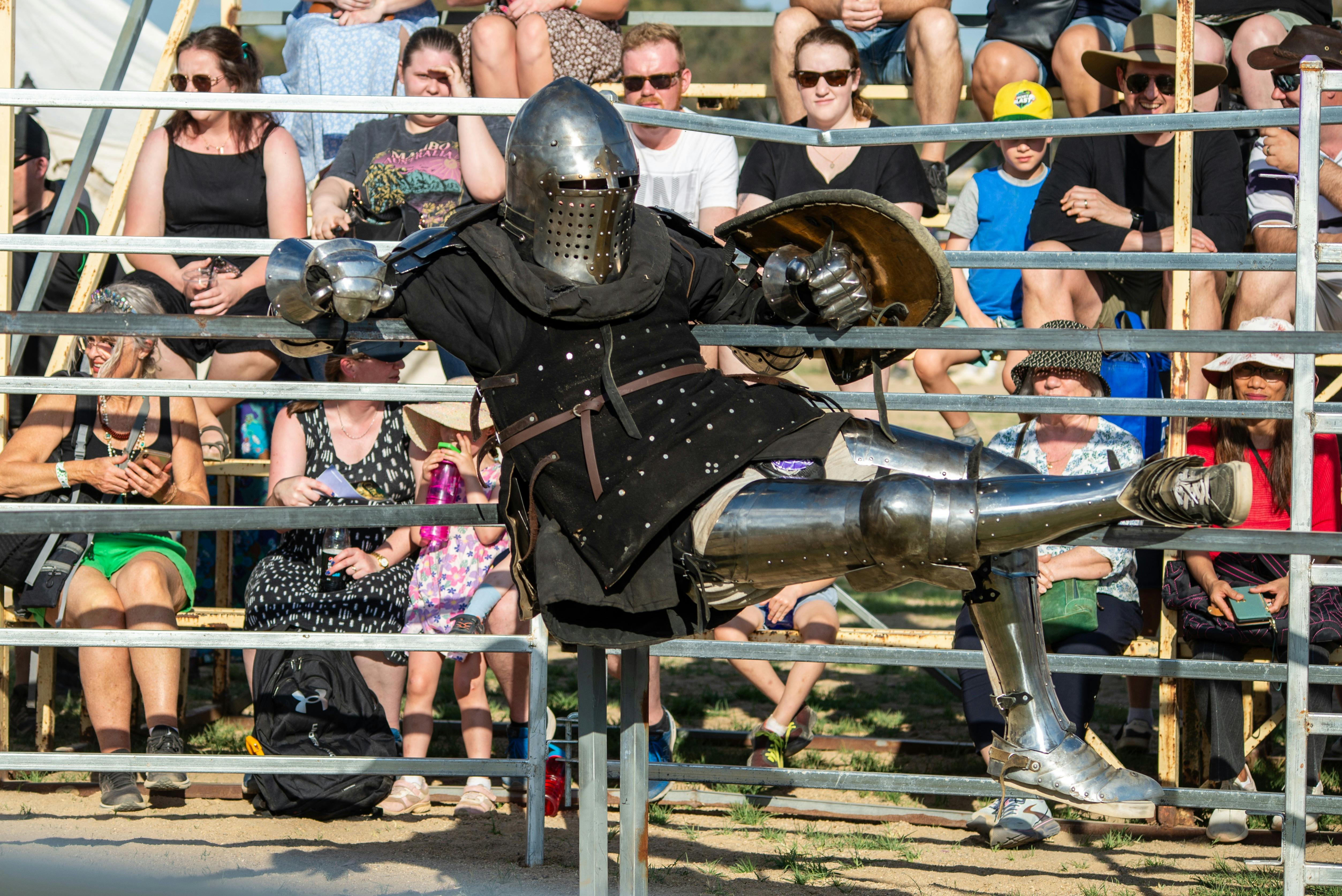Medieval re-enactor dressed as a knight in armour hangs on fence rails in front of people seated.