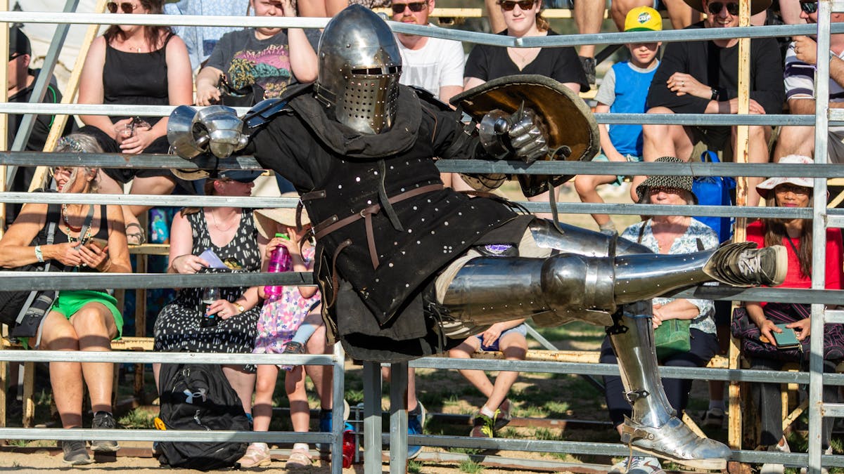 Medieval re-enactor dressed as a knight in armour hangs on fence rails in front of people seated.