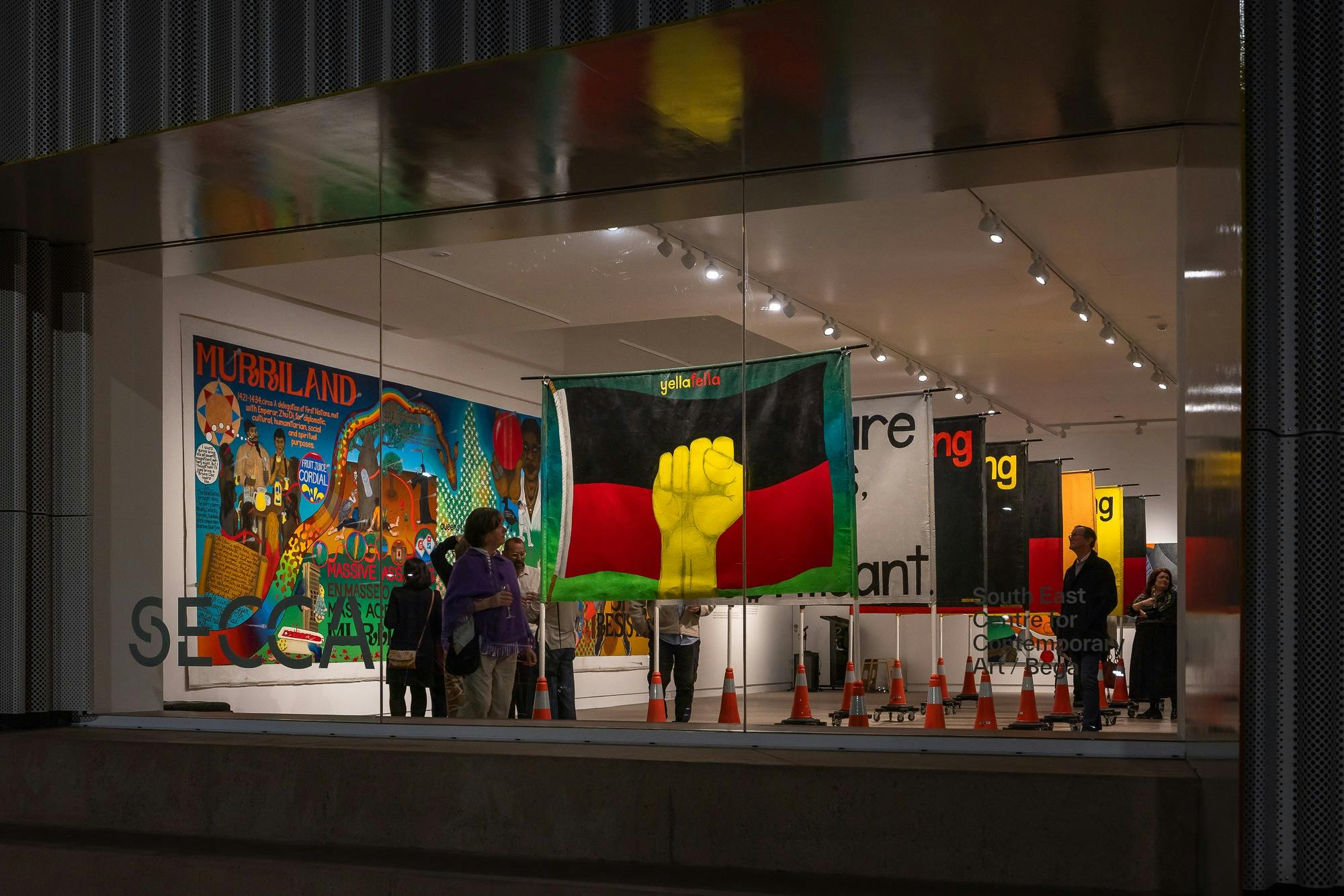 大規模なアボリジニの旗をモチーフにしたアートワークのバナーが表示されているギャラリーの外の窓の夜景。