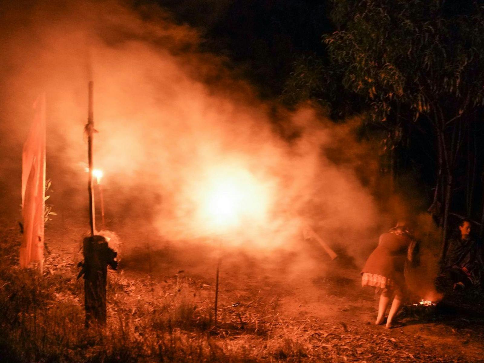 Orange and yellow smudged smoke in the background. A woman bends down to acknowledge the land.