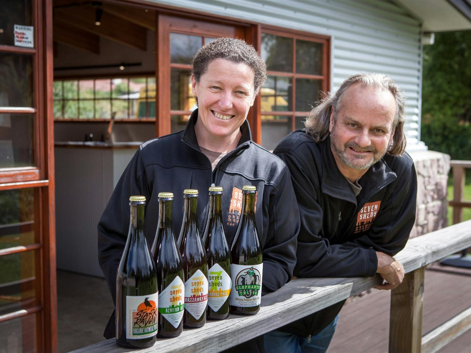 Owners Catherine & Willie leaning on a railing with five bottles of Seven Sheds beer.