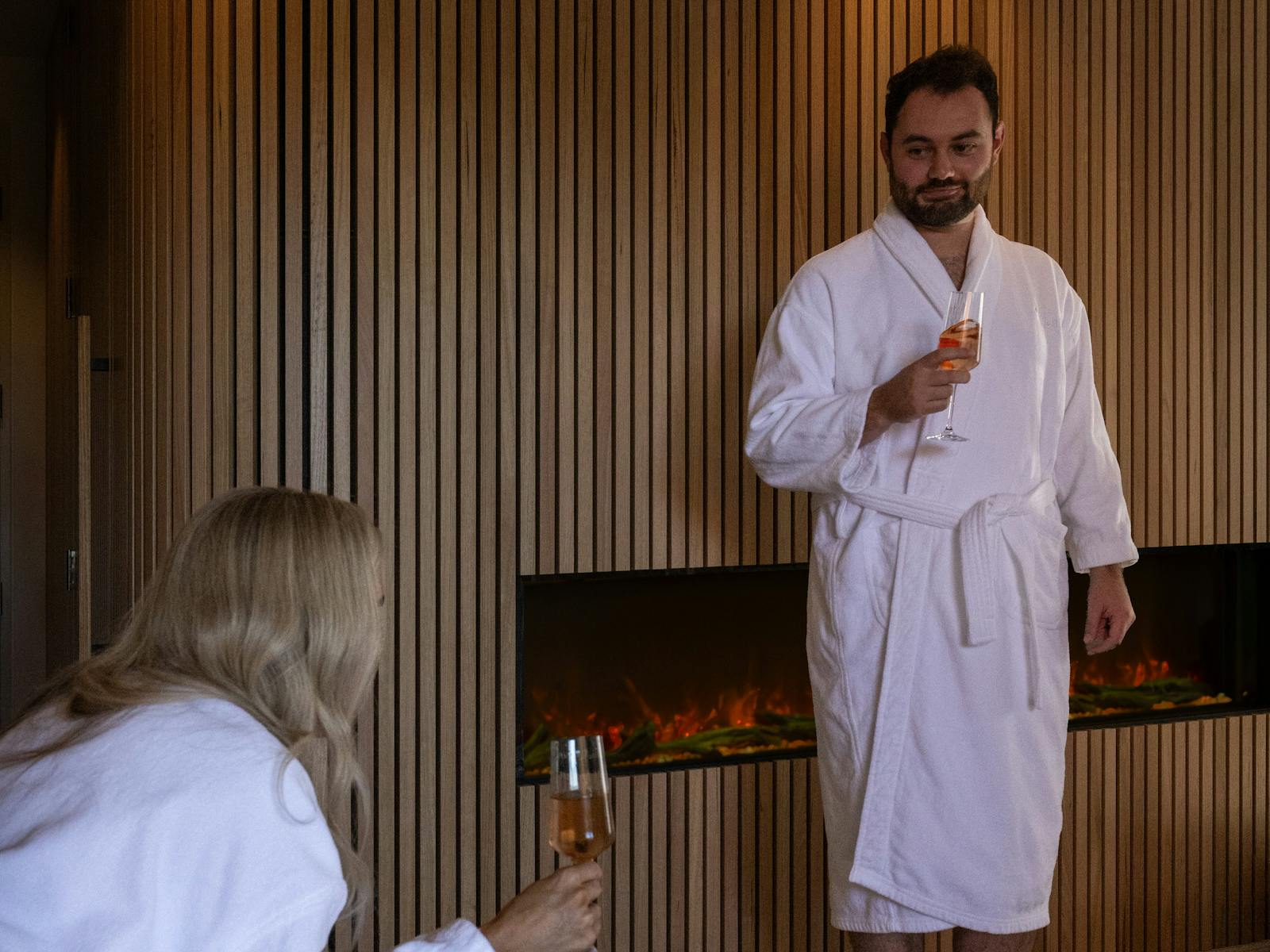 A man enjoying a glass of wine by the fireplace looking at a woman, they are wearing white robes