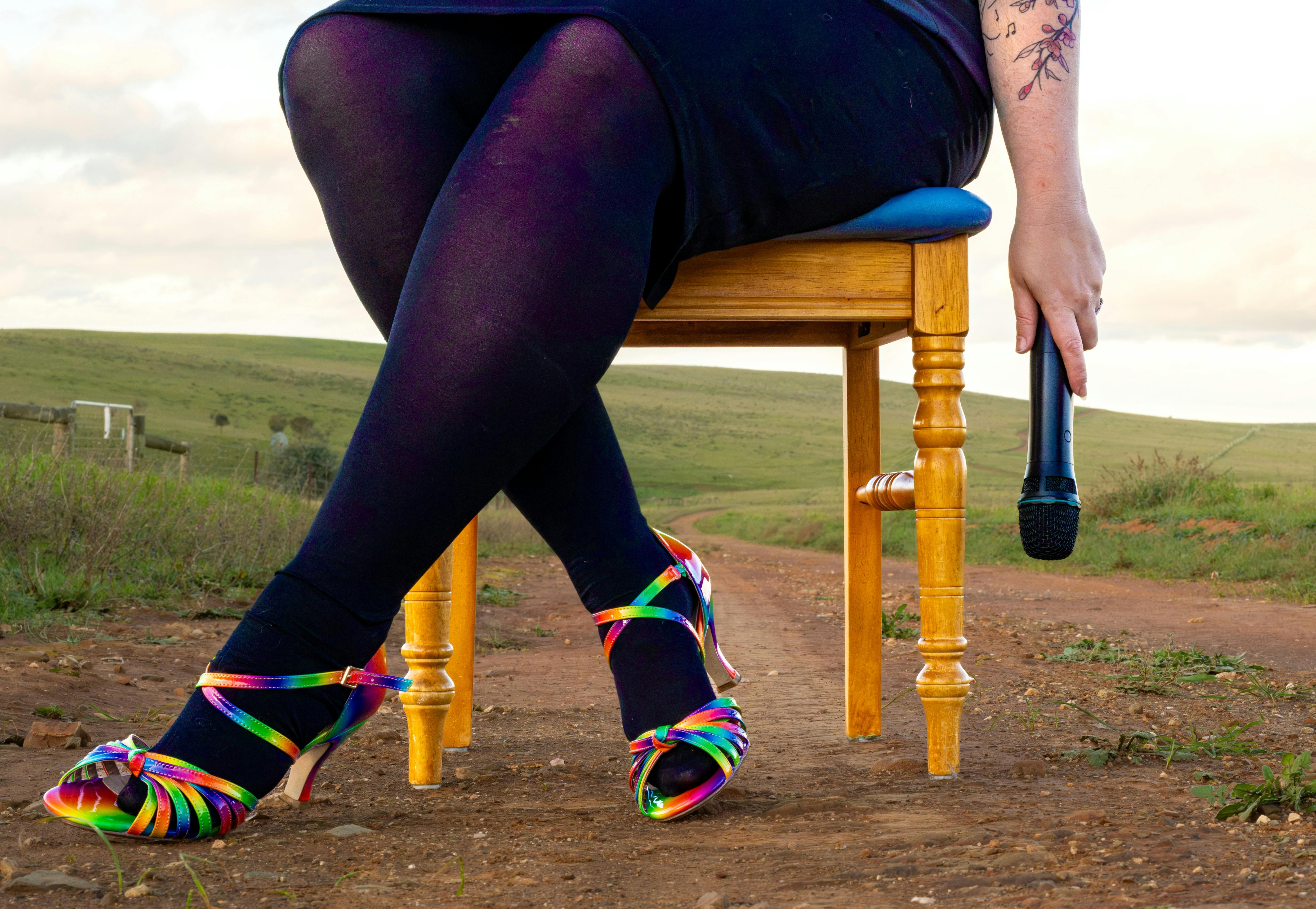 A woman's legs and feet, wearing rainbow shoes. She holds a microphone in a dangling hand.