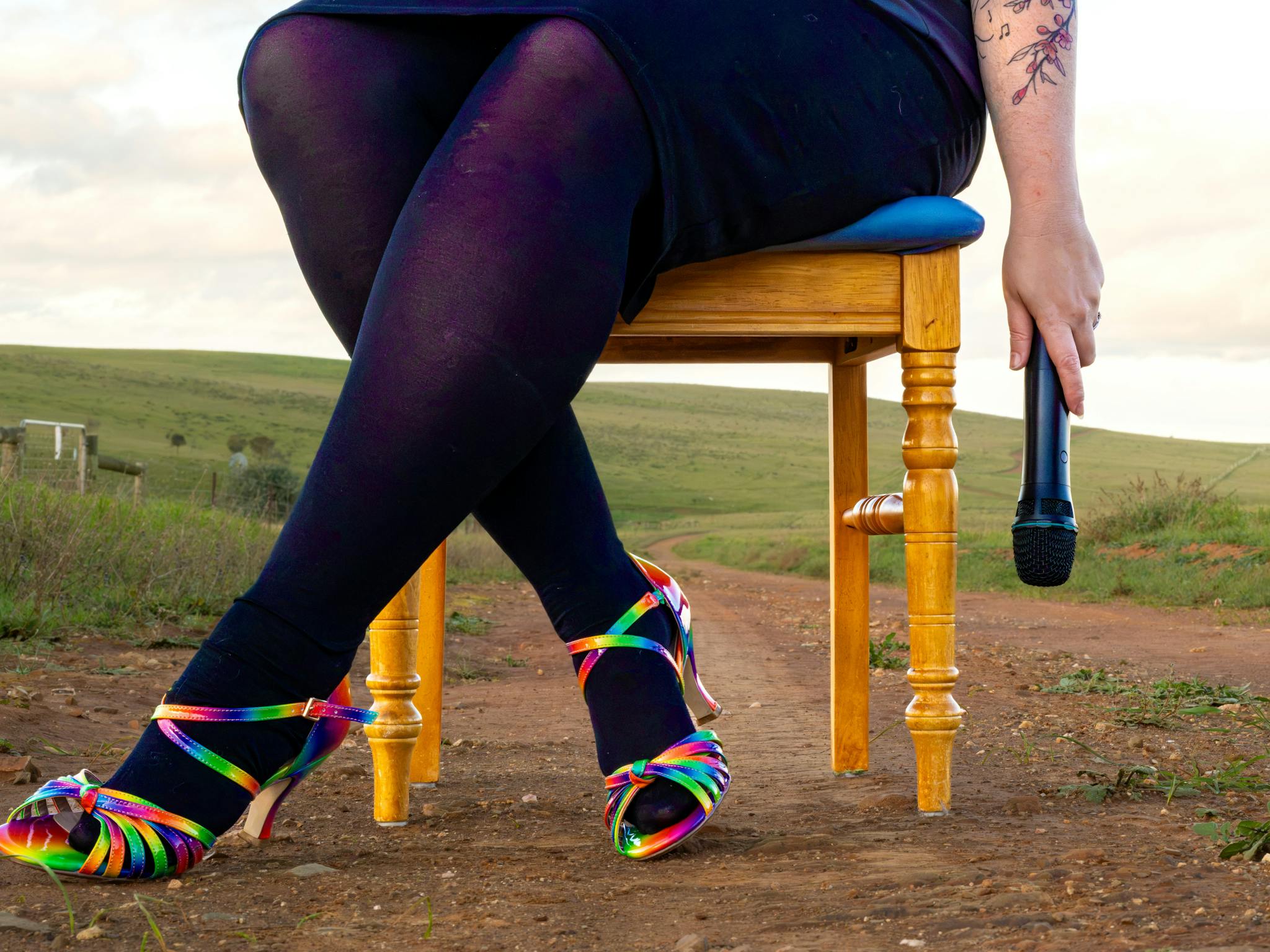 A woman's legs and feet, wearing rainbow shoes. She holds a microphone in a dangling hand.