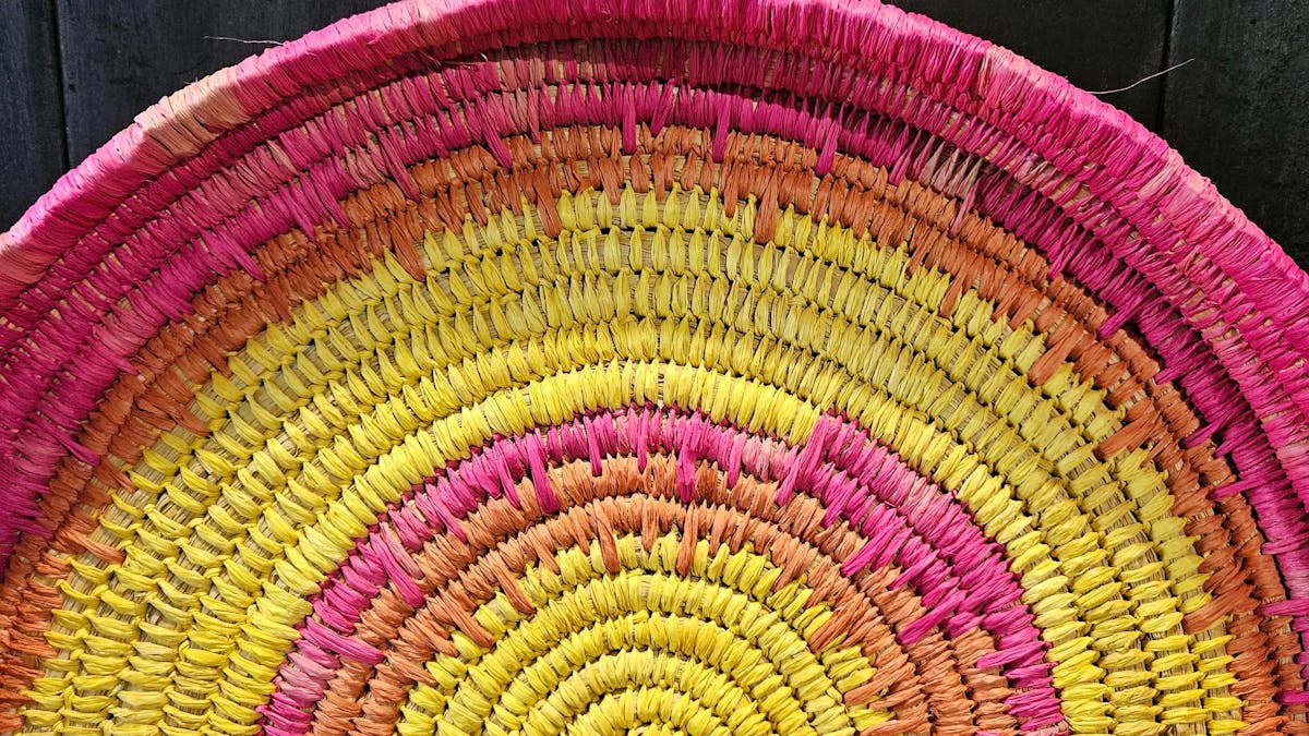 A hand-woven basket in vibrant yellows and pinks against a black wooden wall.