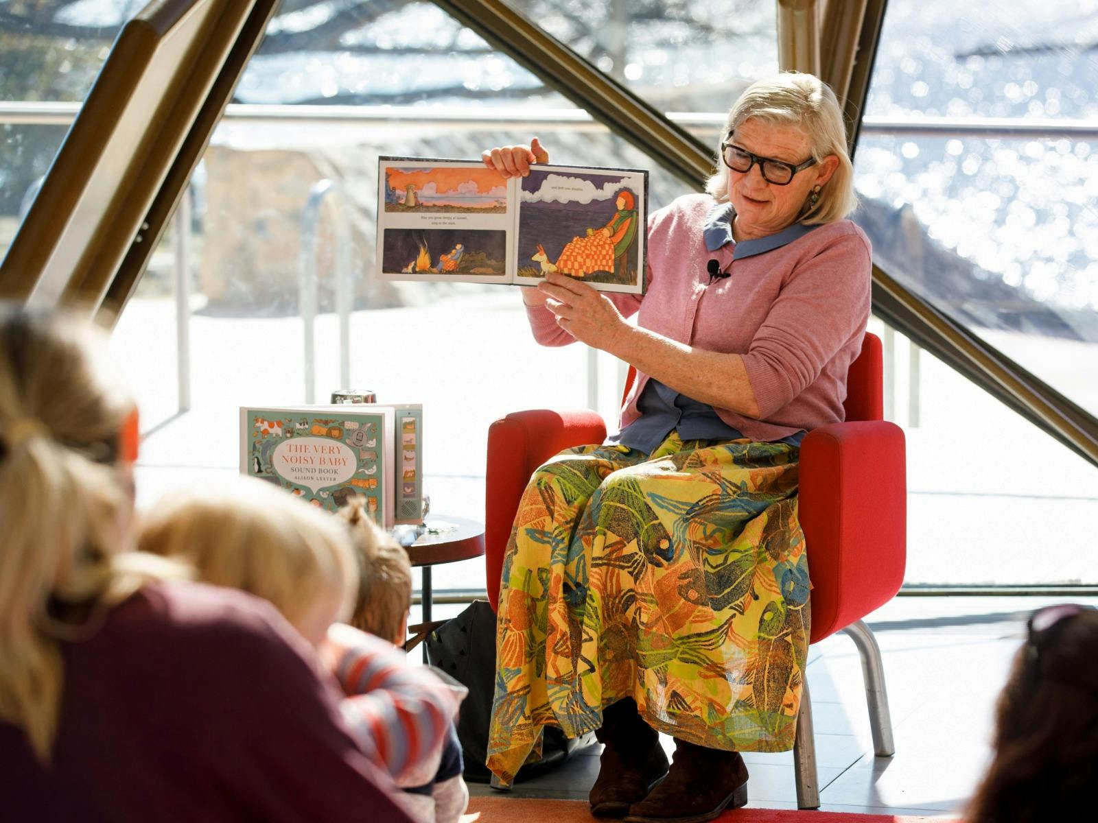 Woman sitting on a chair holding a book reading to a group of children.