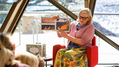 Woman sitting on a chair holding a book reading to a group of children.