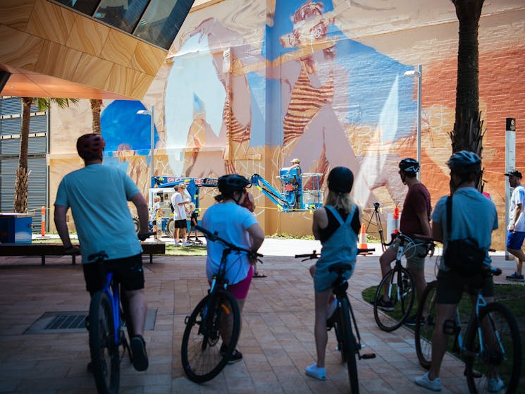 People on bikes watching a massive mural being painted on the side of a city building