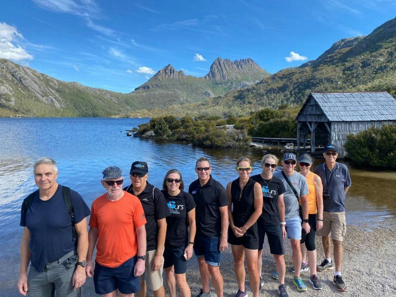 Group of people standing in a row at Dove Lake with old boat shed and Cradle Mountain in background.