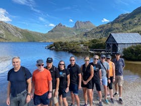 Group of people standing in a row at Dove Lake with old boat shed and Cradle Mountain in background.