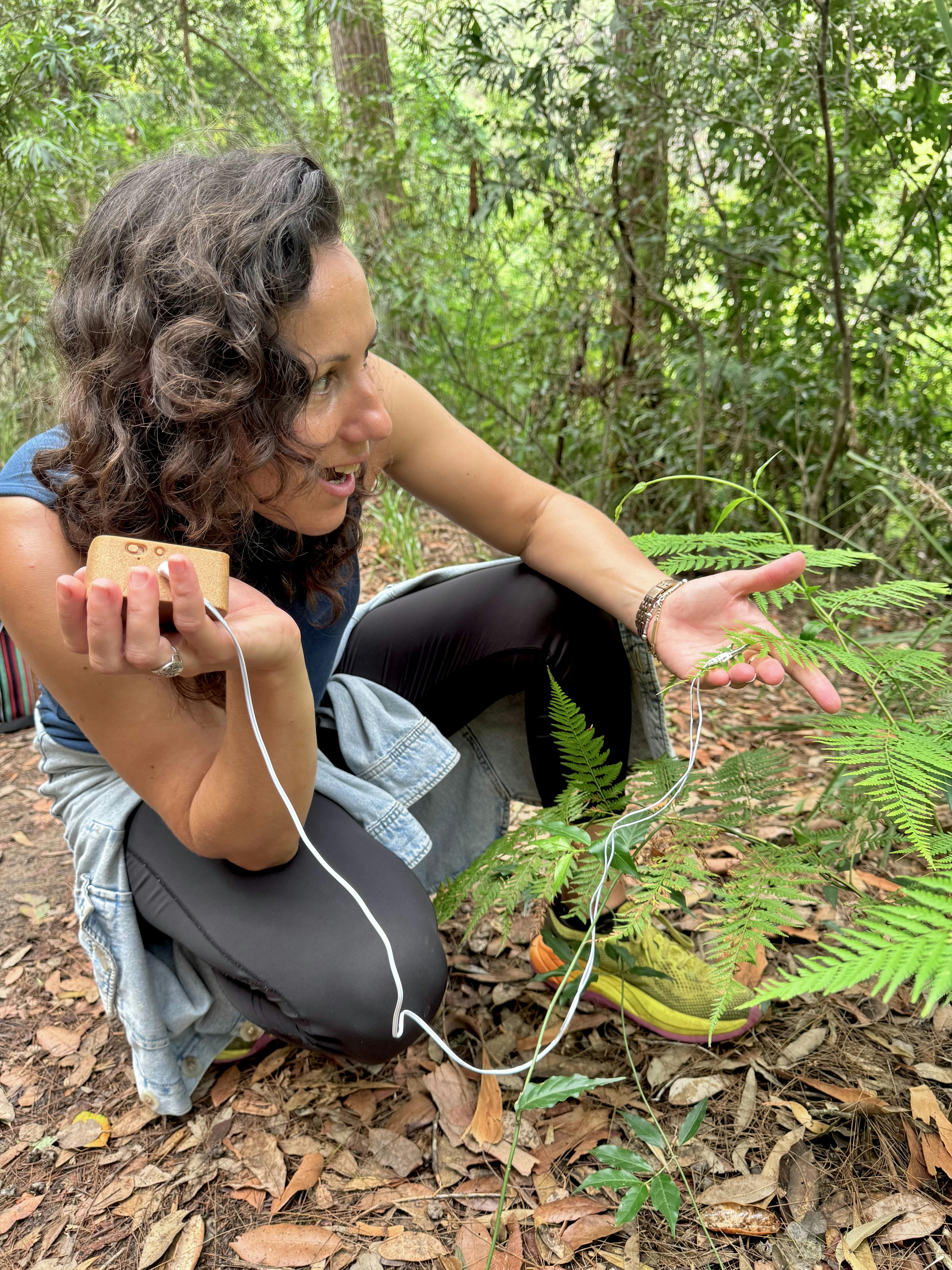 A woman holds and listens to a device connects to a fern via electrodes