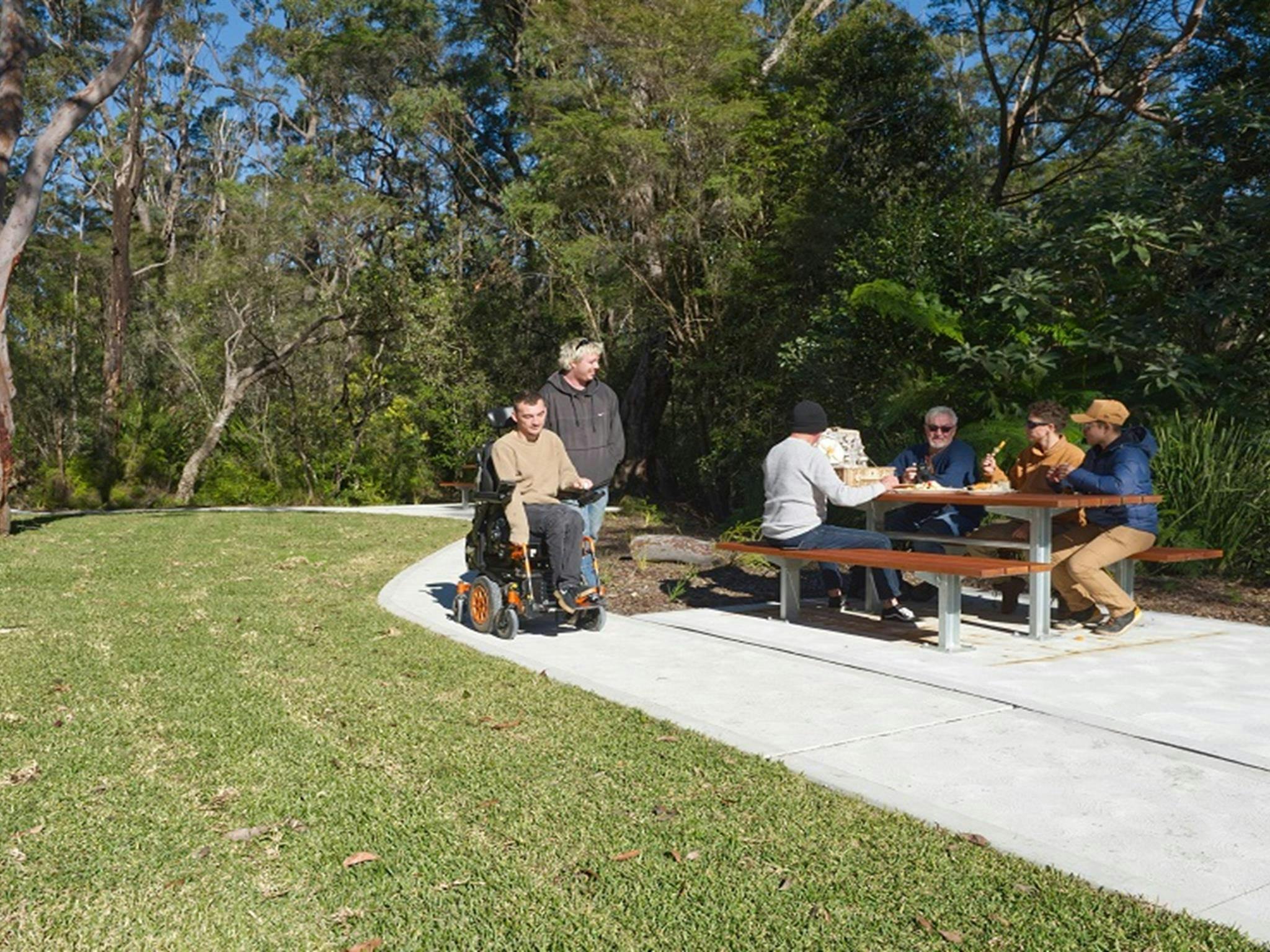 People share lunch at a picnic table, as a man in a wheelchair and his friend pass by, Somersby