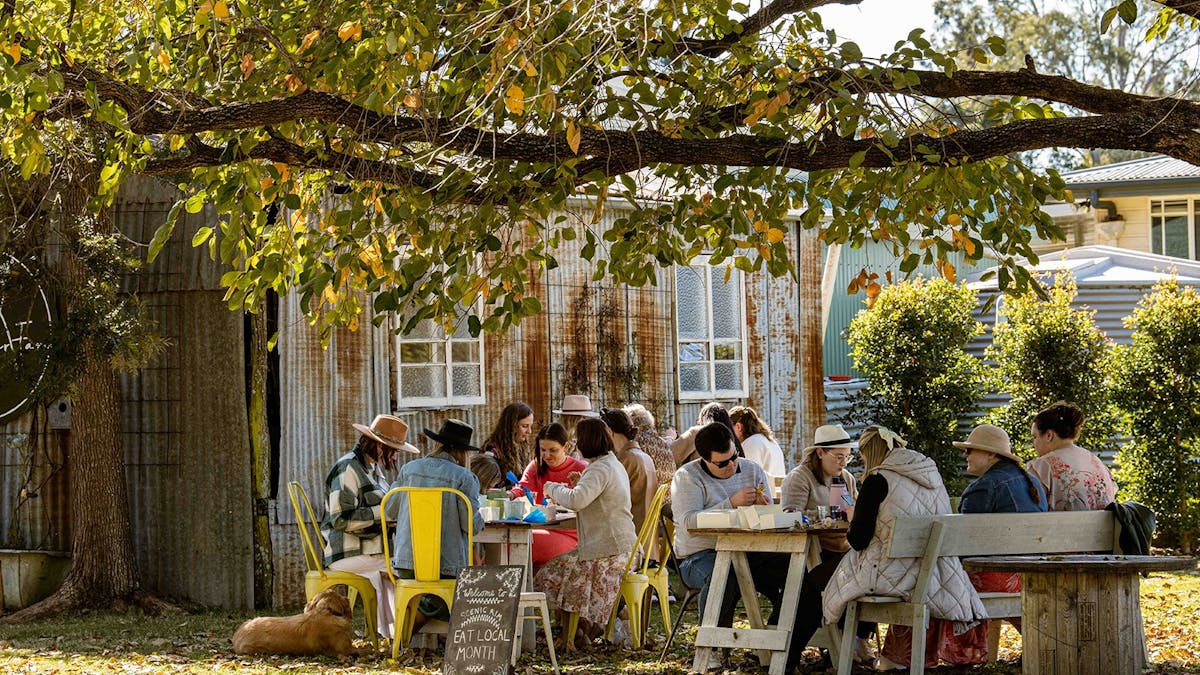 Groups of friends sit around tables underneath a shady tree decorating cupcakes together.