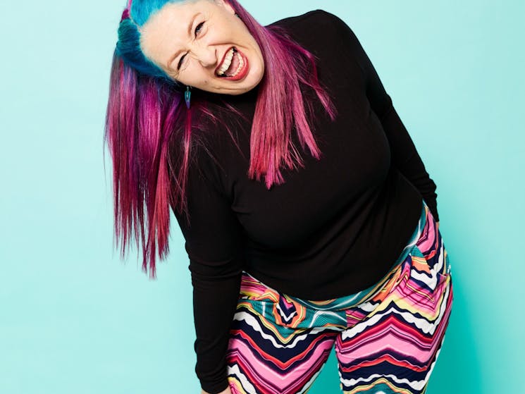 Kelly Mac leans to one side and laughs. She is a middle-aged Caucasian woman with pink and blue hair