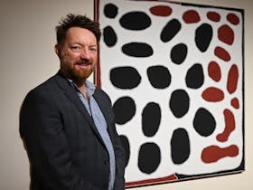 A man with short hair stands side on to a large artwork. He has facial hair and is wearing a suit