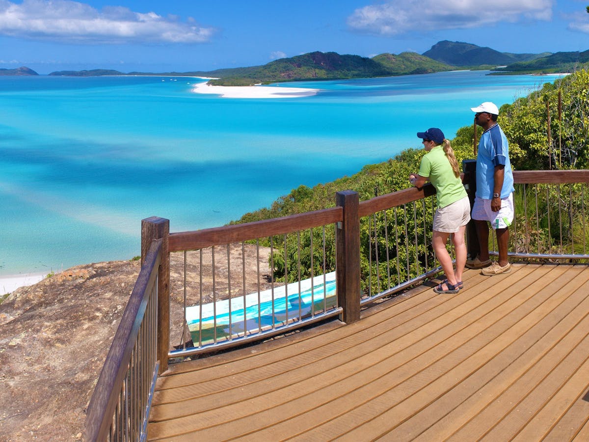 A couple standing on a wooden lookout platform looking out over turquoise water, white sand & island