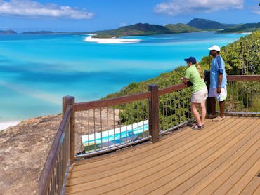 A couple standing on a wooden lookout platform looking out over turquoise water, white sand & island