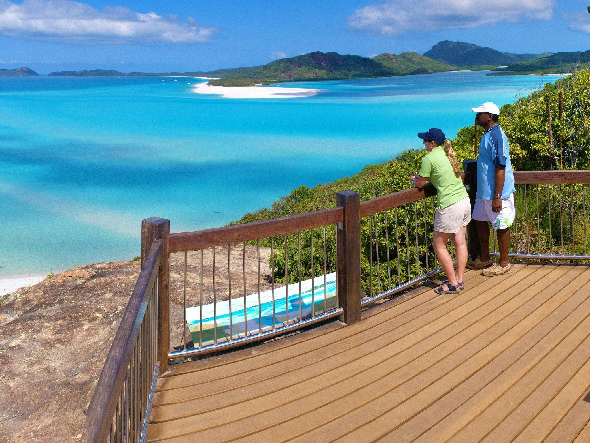 A couple standing on a wooden lookout platform looking out over turquoise water, white sand & island A couple standing on a wooden lookout platform looking out over turquoise water, white sand & island