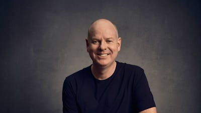 Comedian Tom Gleeson sitting on a stool, smiling