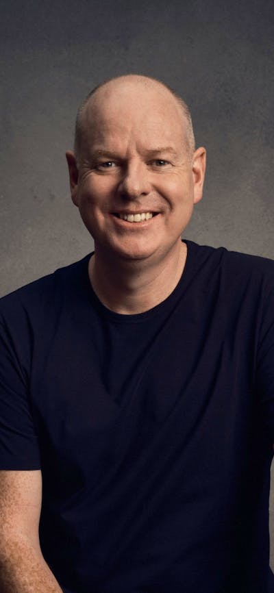 Comedian Tom Gleeson sitting on a stool, smiling