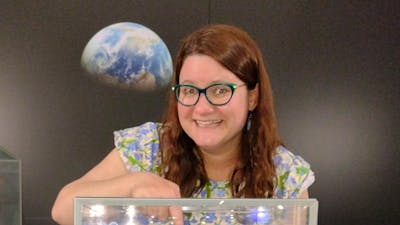 A woman touches a small rock in a cabinet with an image of the Moon and Earth behind her.