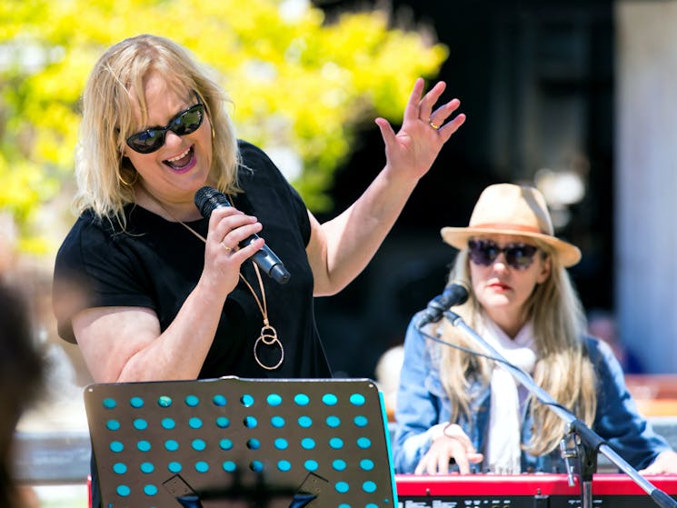 A woman with blonde hair and a black top is singing into a microphone with a music stand in front