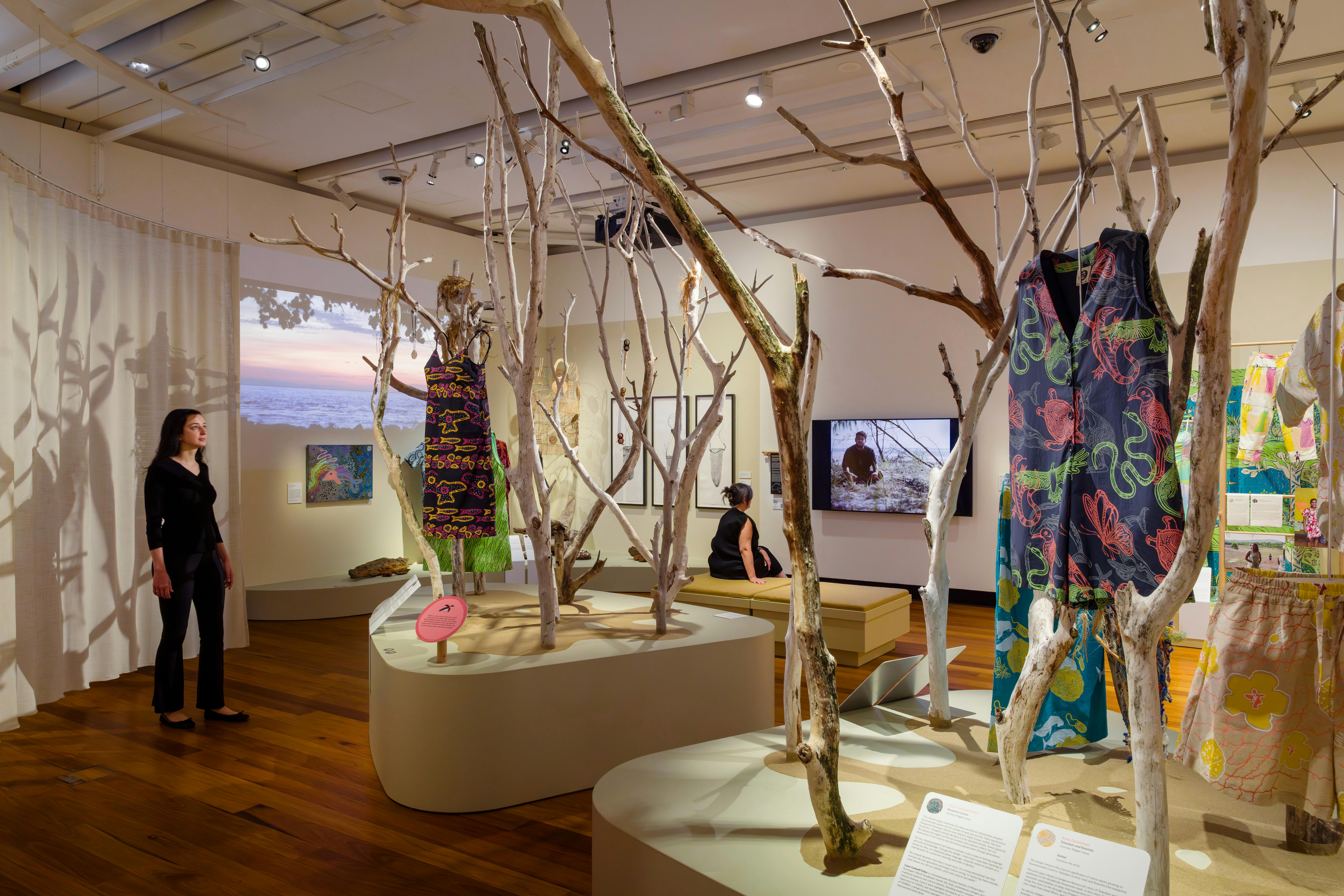 Stories You Wear exhibition, visitors in the space where branches are upright & clothing pieces hang