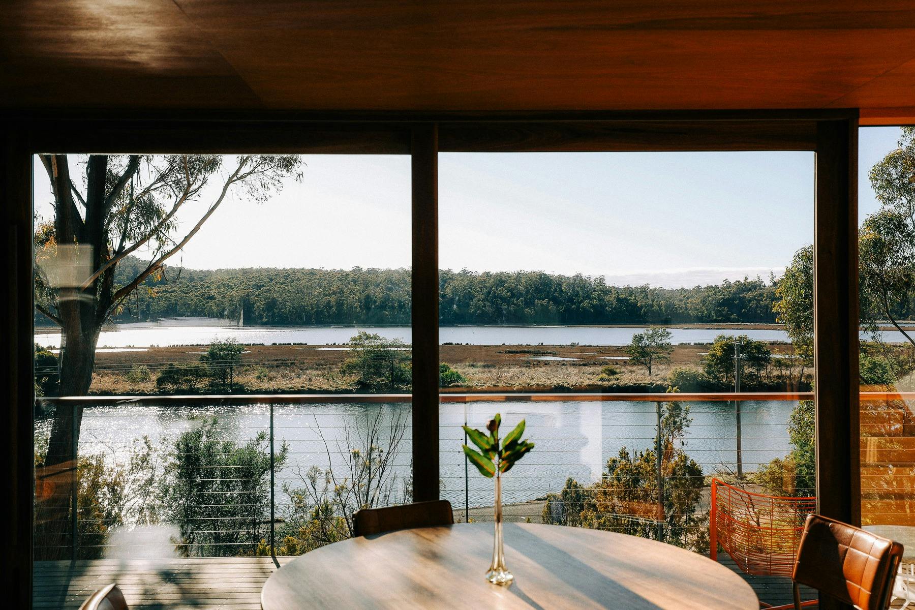 A round dining table with floor to ceiling windows behind overlooks a river.