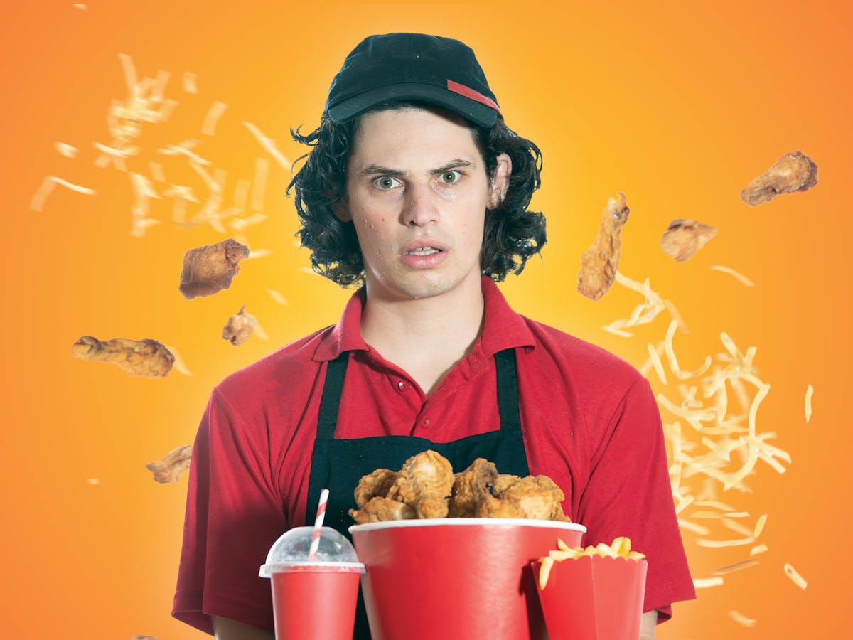A young man in a fast-food uniform looks bewildered as fried chicken and chips fly around him.