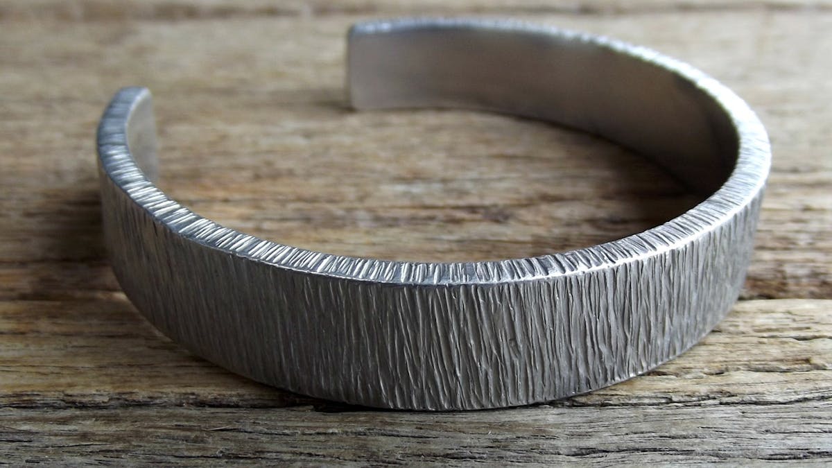 Silver-smithed bracelet with textured pattern displayed on wooden bench.