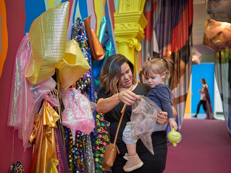 Women with toddler on hip chooses headdress for child to try on from colourful rack of costumes