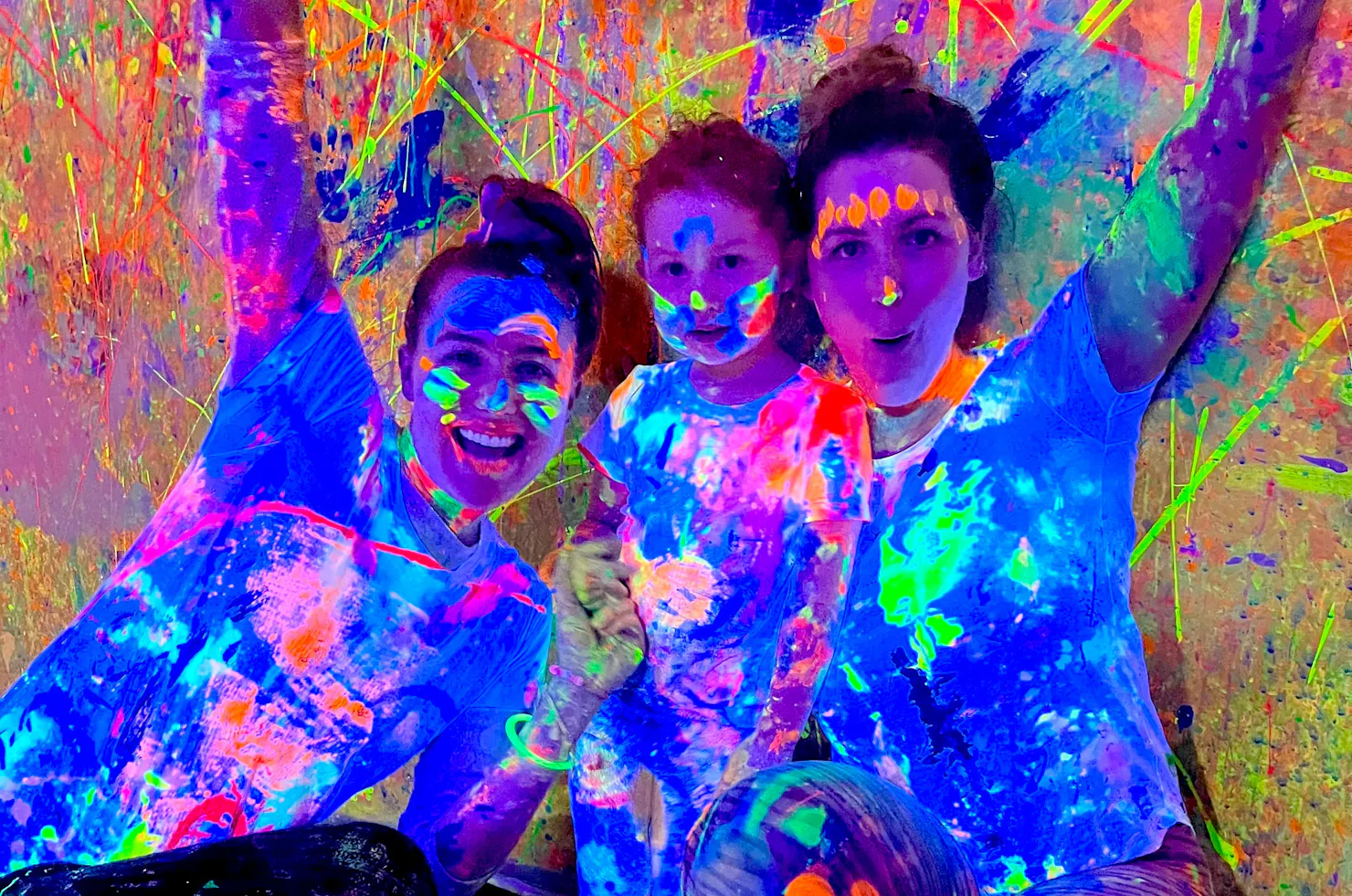 A family of two adults and one child with hands in the air covered in glowing paint at Messy Glow Co