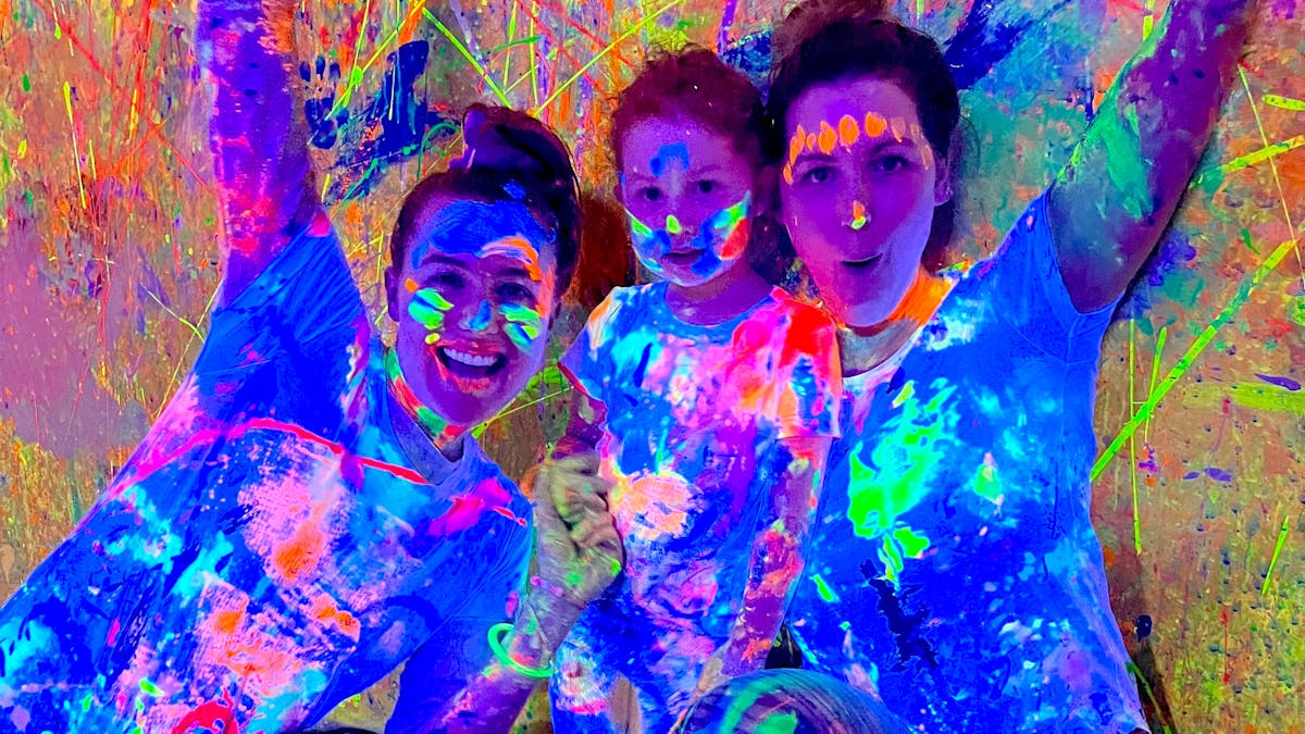 A family of two adults and one child with hands in the air covered in glowing paint at Messy Glow Co