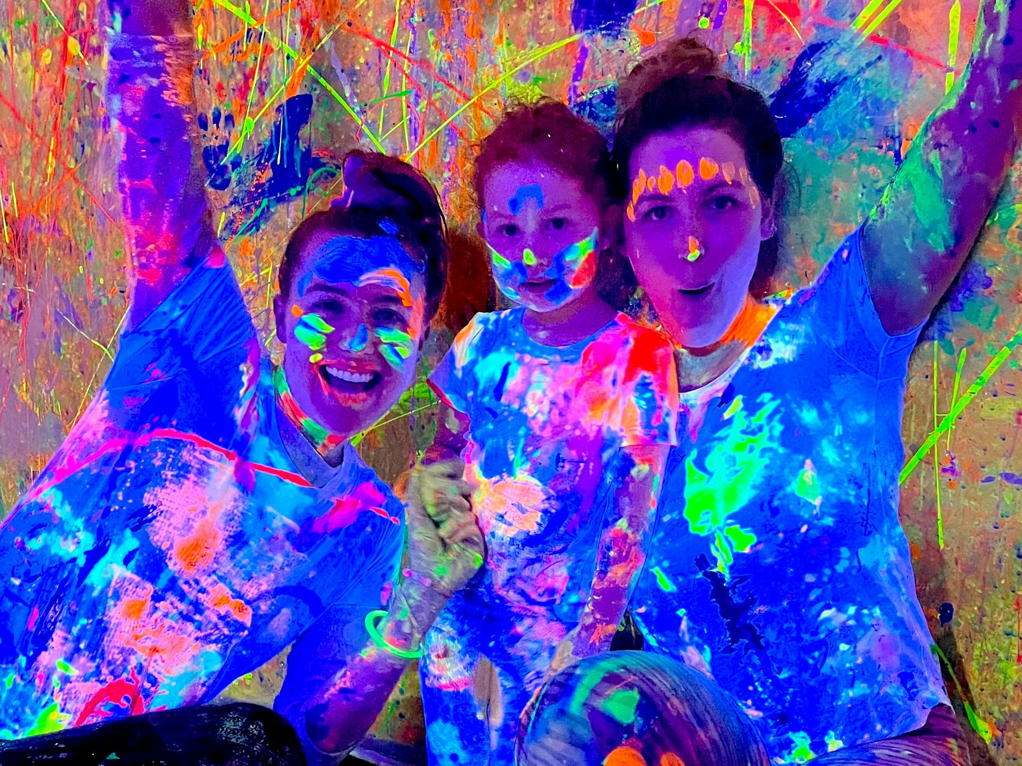 A family of two adults and one child with hands in the air covered in glowing paint at Messy Glow Co A family of two adults and one child with hands in the air covered in glowing paint at Messy Glow Co