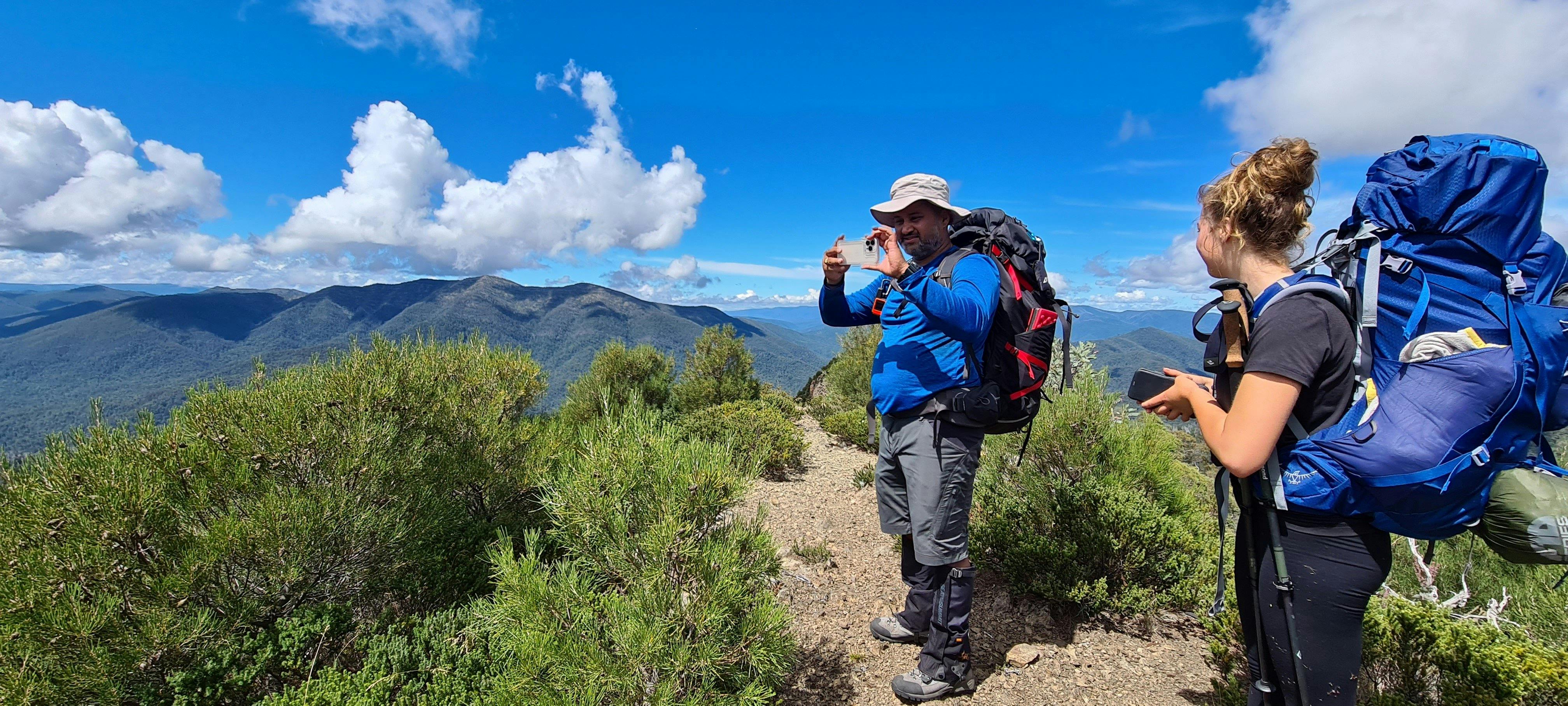A couple of hikers capturing the surrounding mountain views on their phone cameras.