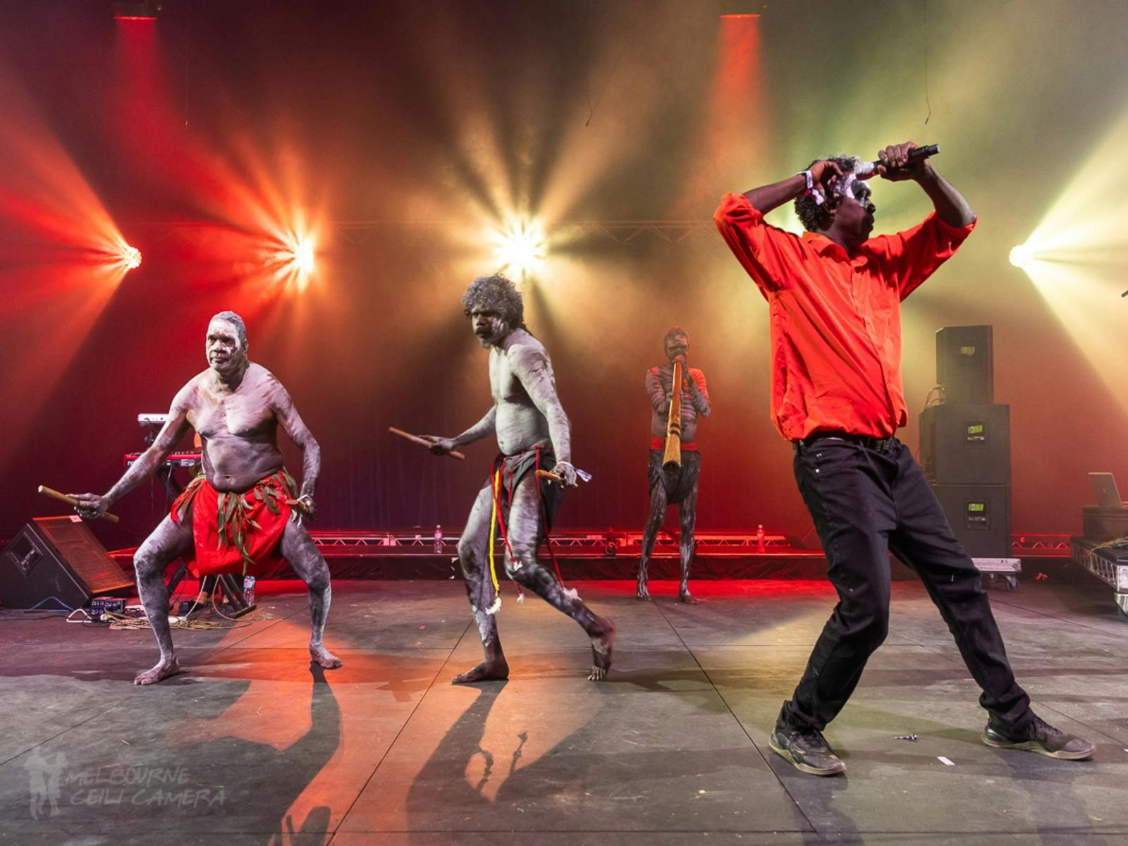 3 members of Yothu Yindi perform. There are 2 dancers in the background in traditional dress