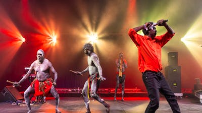 3 members of Yothu Yindi perform. There are 2 dancers in the background in traditional dress