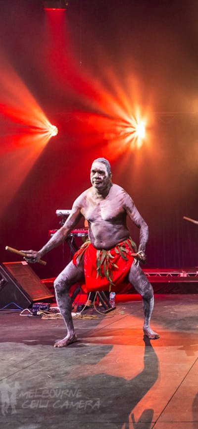 3 members of Yothu Yindi perform. There are 2 dancers in the background in traditional dress