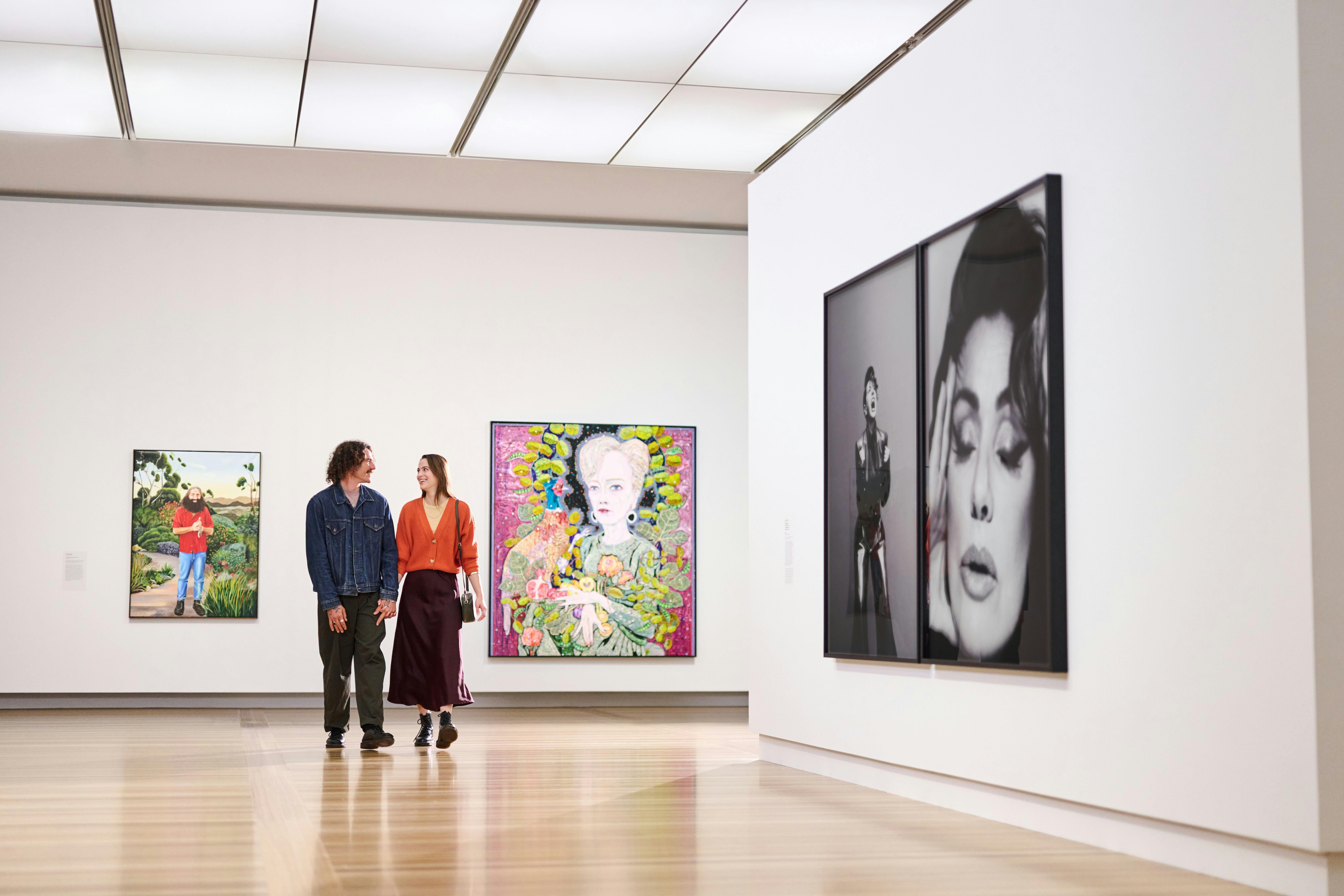 A couple is walking happily through a brightly lit gallery featuring several large portraits.